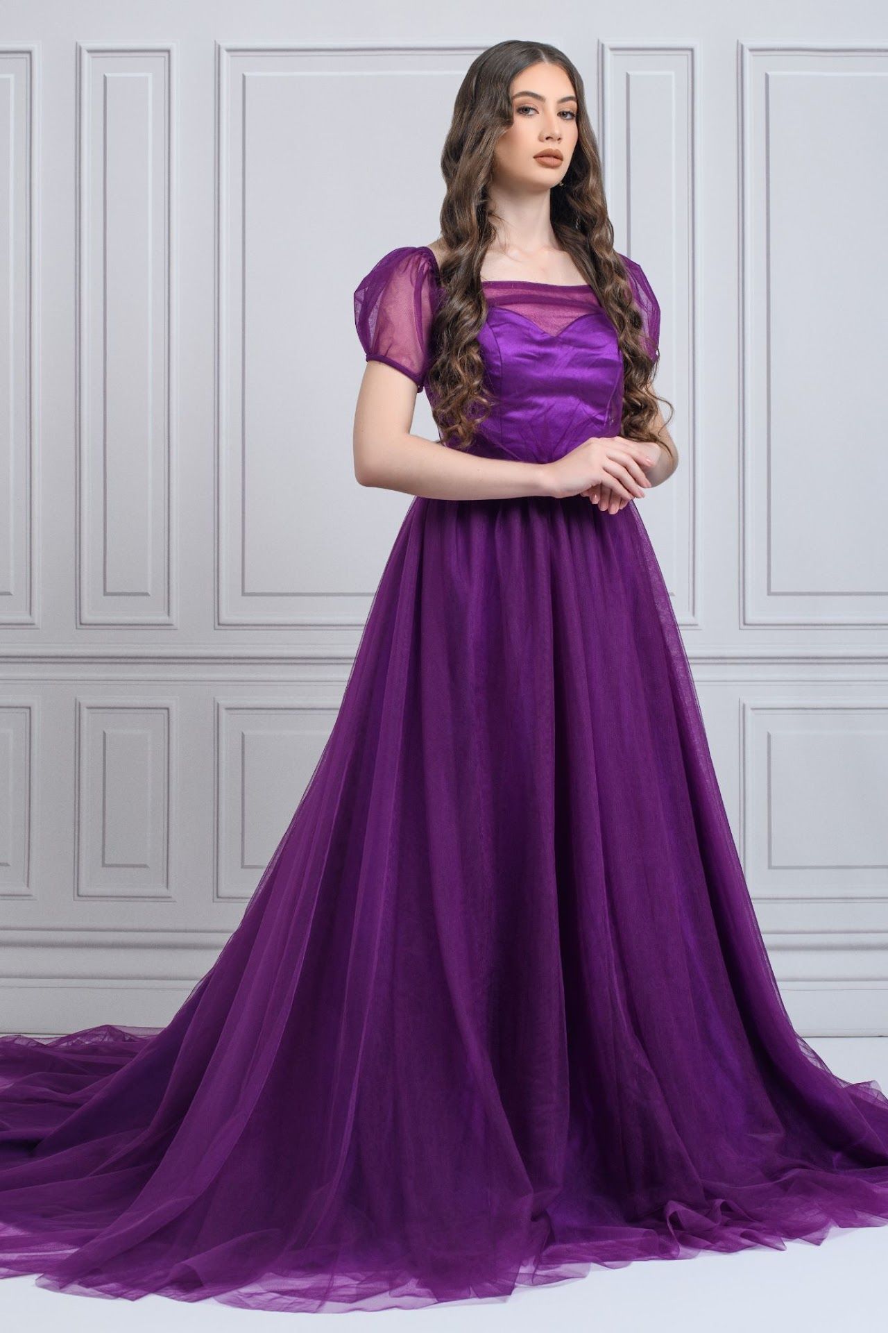 Elegant purple ball gown with puff sleeves and long flowing train against white paneled wall backdrop.