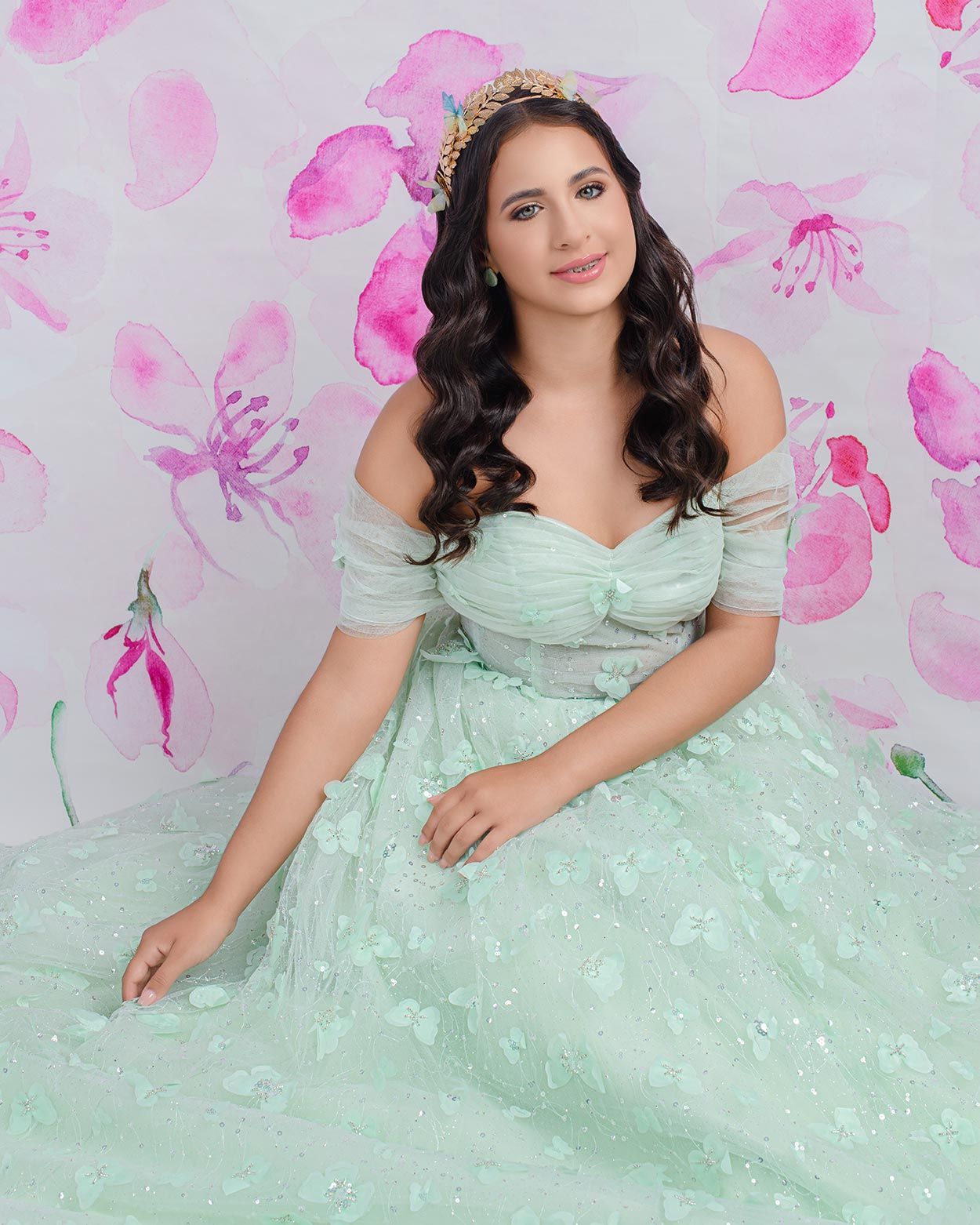 A person in a mint green ball gown with off-shoulder design poses against a pink watercolor floral backdrop.