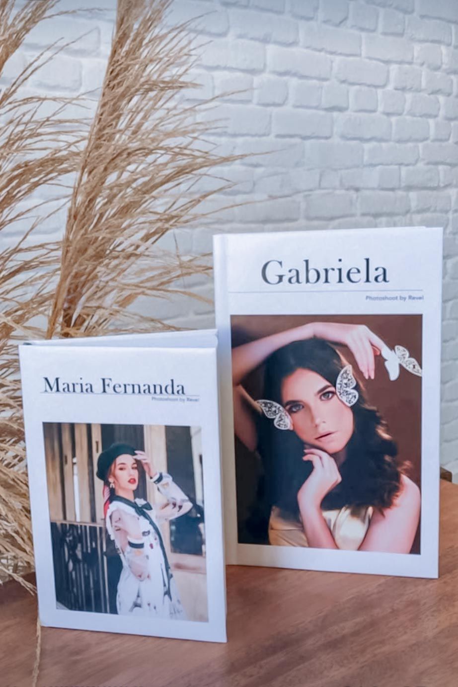 Two fashion magazines or portfolios with decorative dried pampas grass against a white brick wall background.