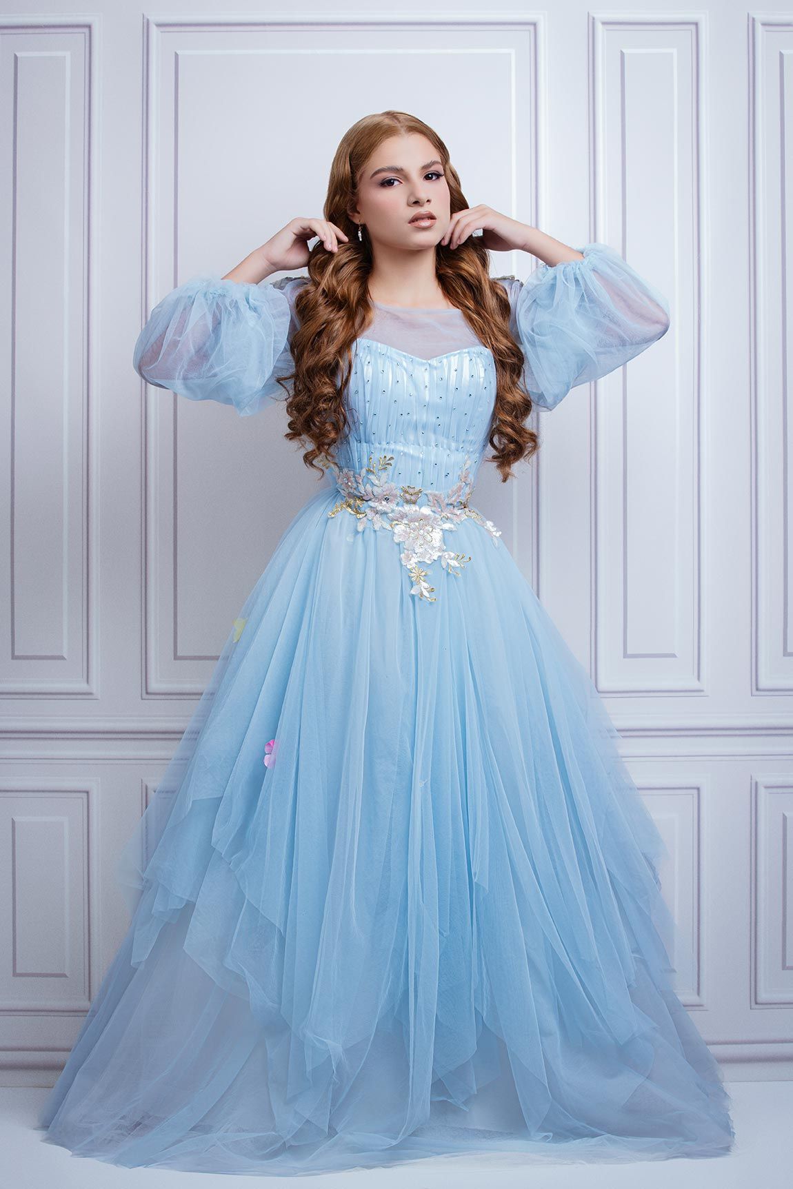 A graceful figure in a flowing light blue tulle ball gown with puff sleeves and delicate floral details against a white paneled wall.