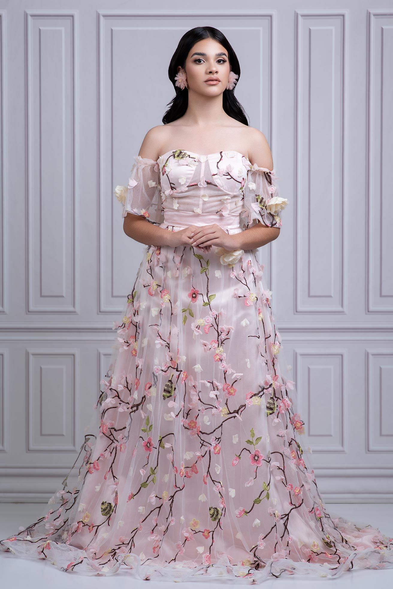 Elegant off-shoulder pink floral ball gown with delicate cherry blossom pattern and sweeping train against grey paneled wall.
