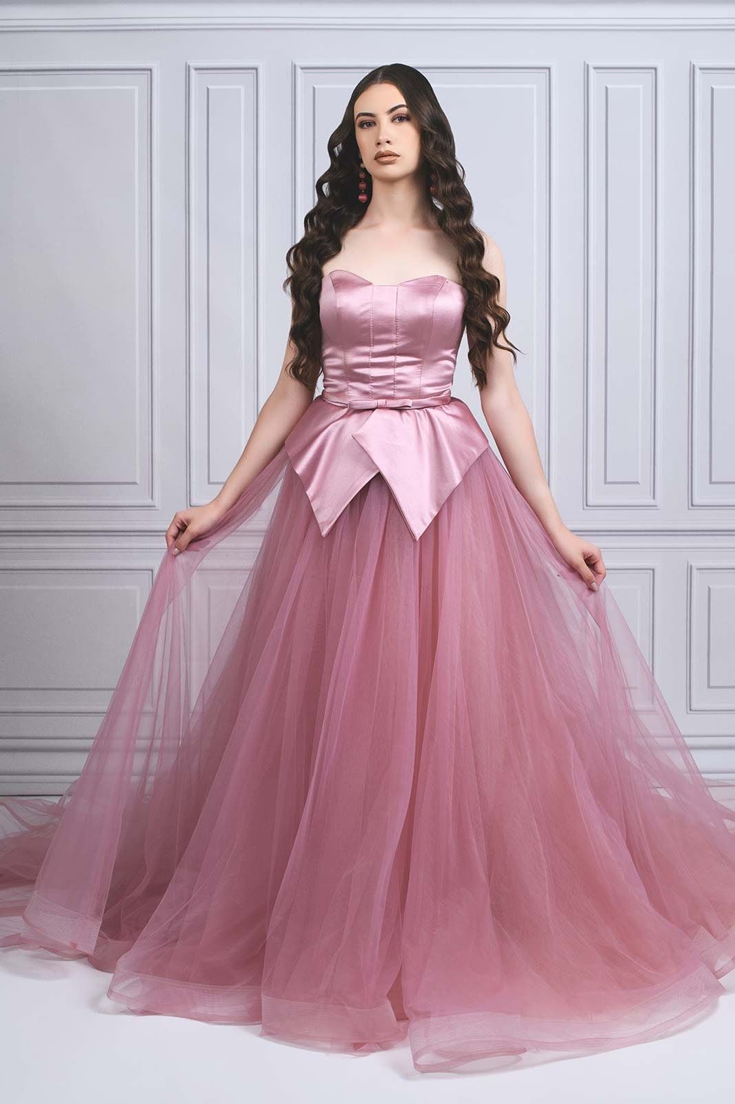 A model in a pink tulle ball gown with satin bodice and bow detail poses against a classic white wall backdrop.