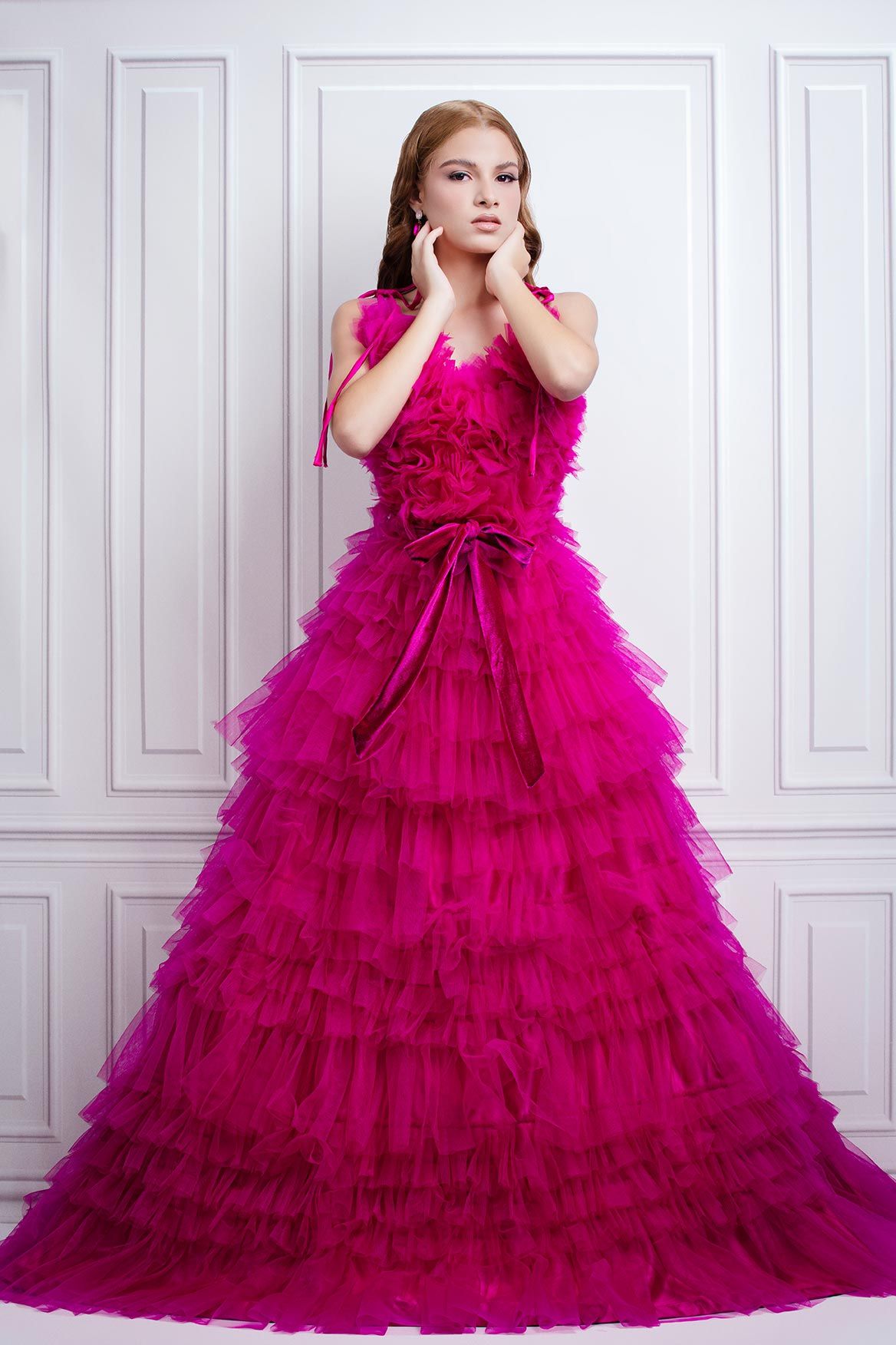 Stunning magenta layered tulle ball gown with ruffled tiers and bow detail photographed against white paneled wall.