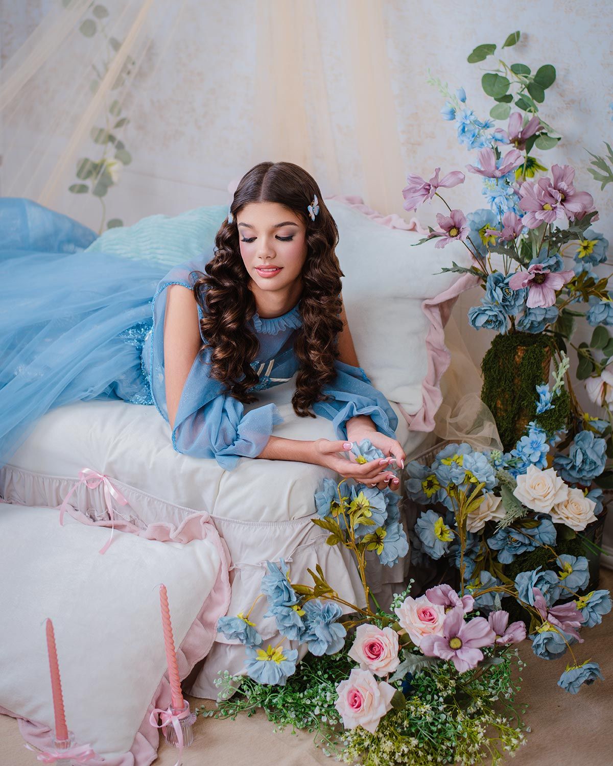 A dreamy boudoir scene with a flowing blue dress, soft pillows, and romantic floral arrangements of roses and delphiniums.
