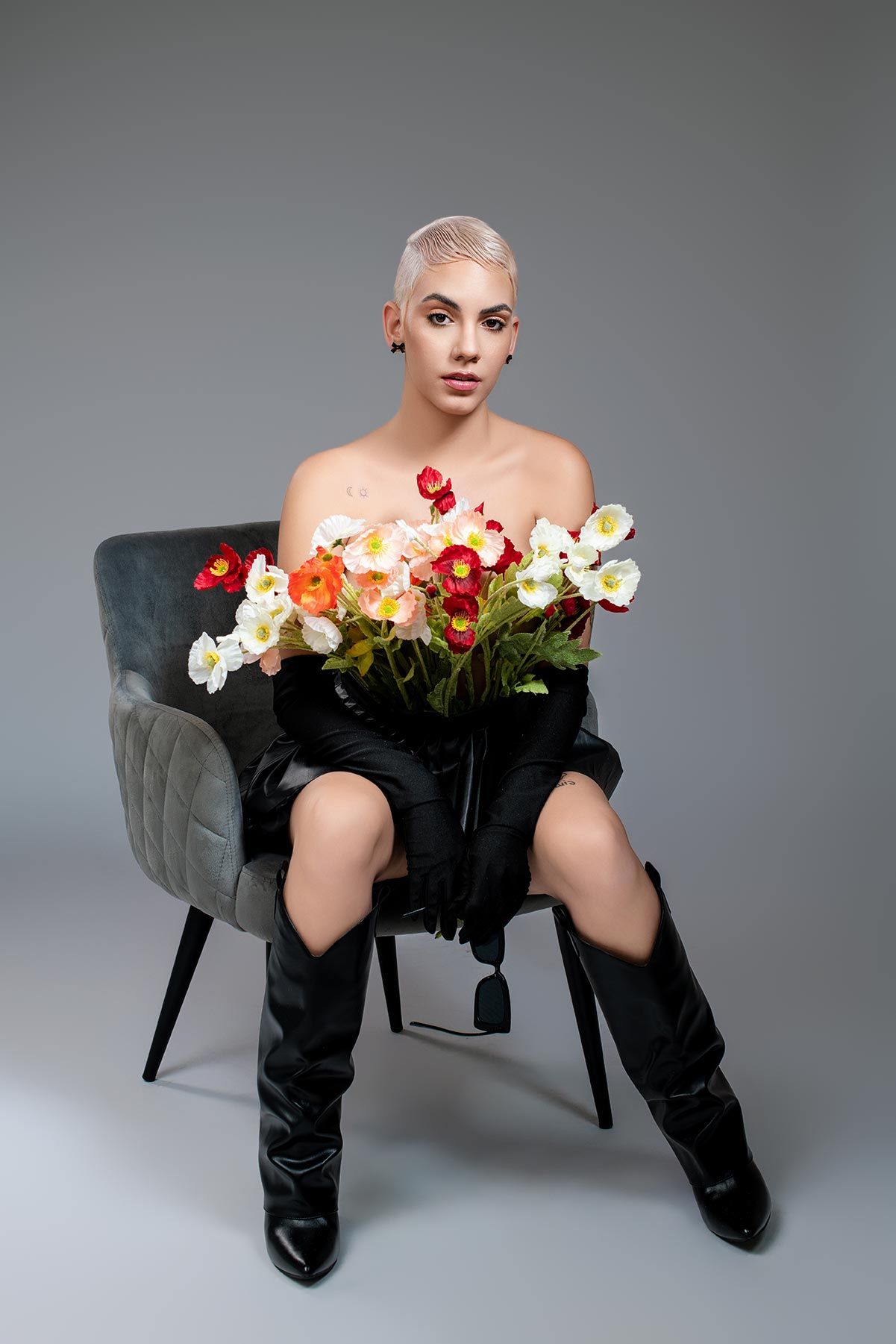 Artistic fashion portrait with colorful flowers on grey velvet chair against neutral background.