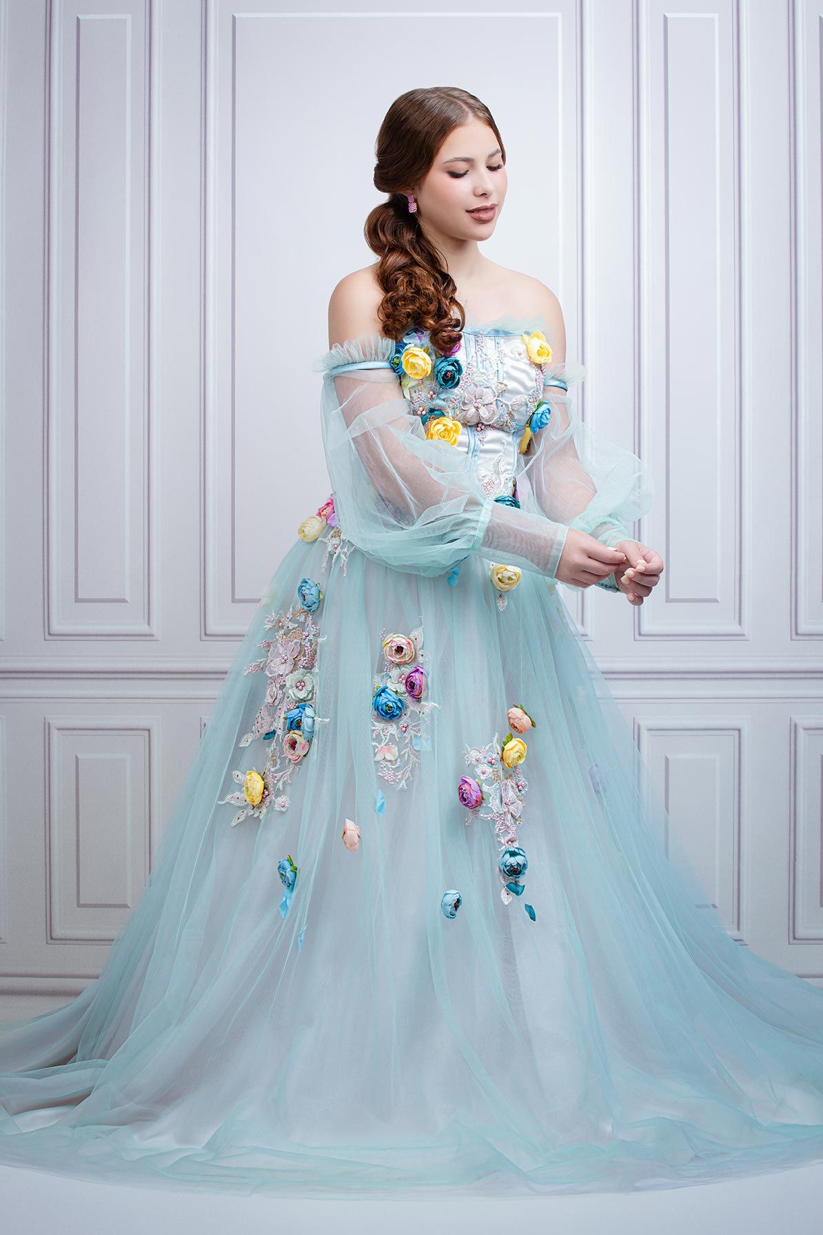 Ethereal light blue ball gown with floral appliques and sheer sleeves photographed against white paneled wall.