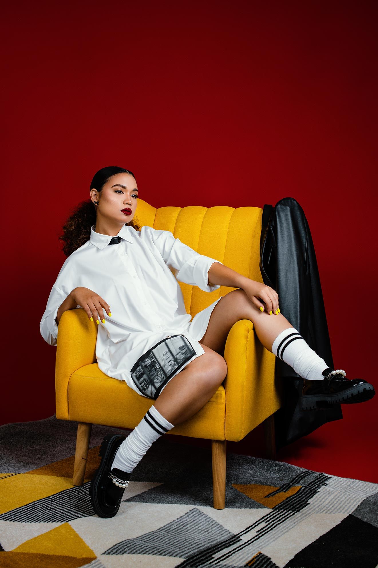 Fashion photo with person lounging in bright yellow armchair against red wall wearing white shirt and striped shorts.
