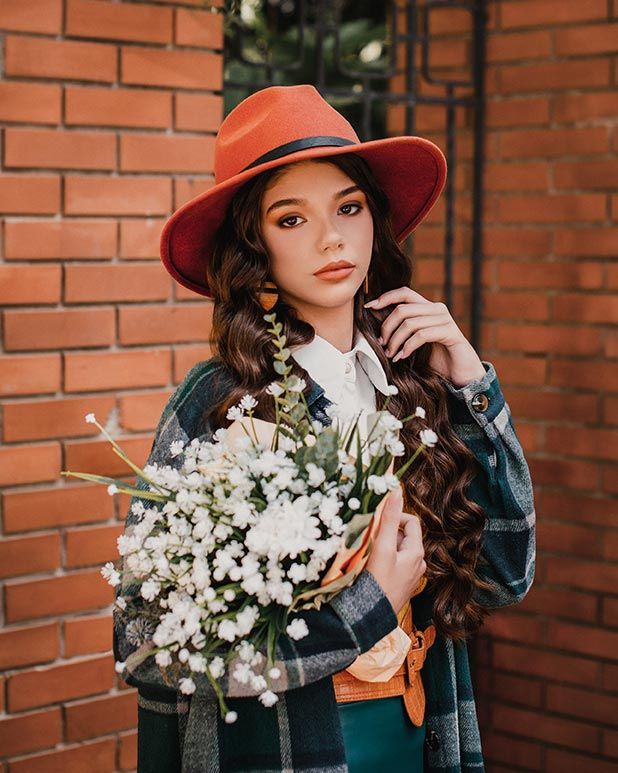 Someone in a denim jacket and red hat holds white baby's breath flowers while posing against a brick wall backdrop.