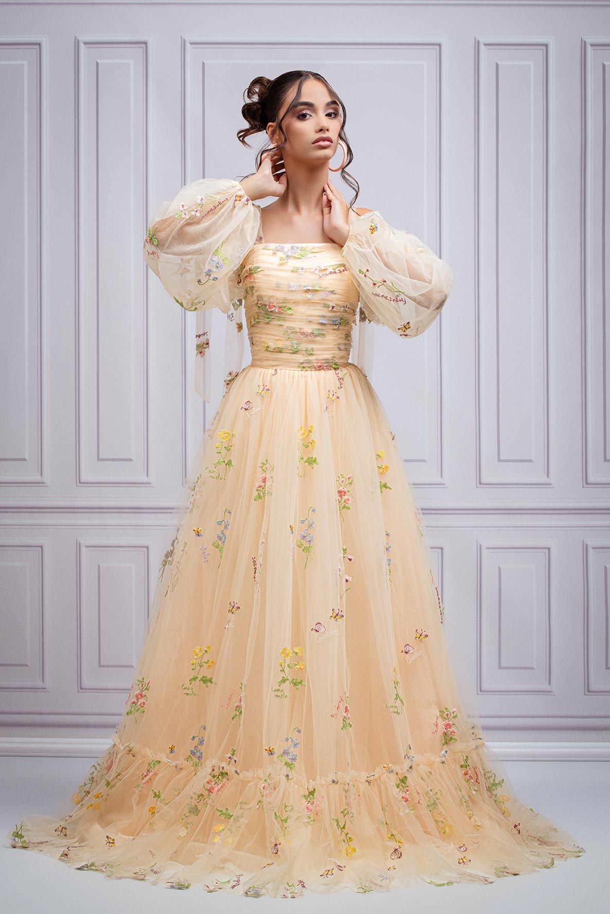 Romantic peach ball gown with puffy sleeves and delicate floral embroidery on tulle against white wall backdrop.