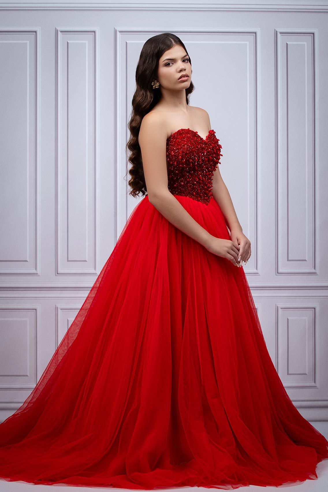 A stunning red ball gown with beaded sweetheart bodice and flowing tulle skirt photographed against a white paneled wall.