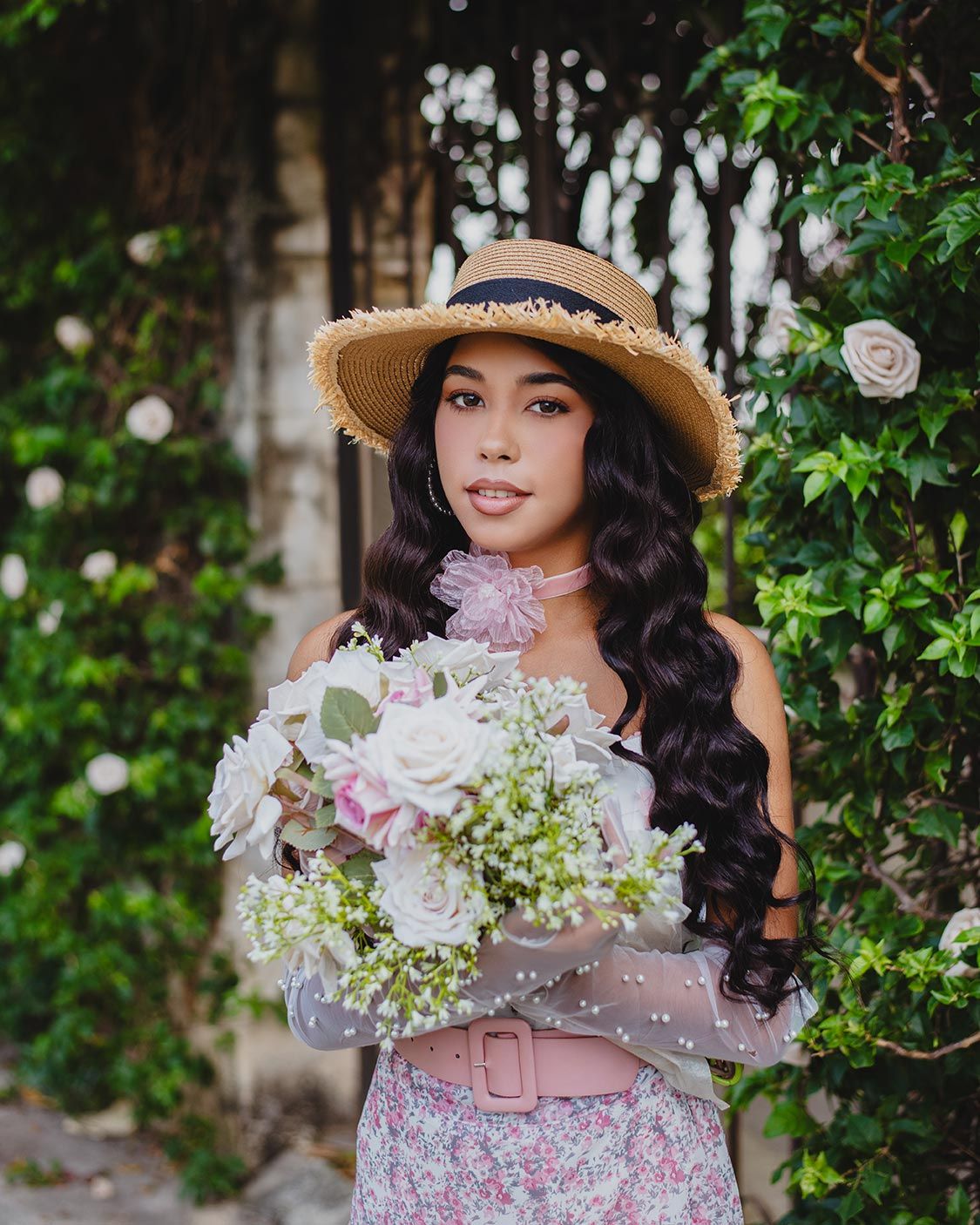 A person in a straw hat and floral dress holds a beautiful bouquet of white and pink flowers against a green garden backdrop.