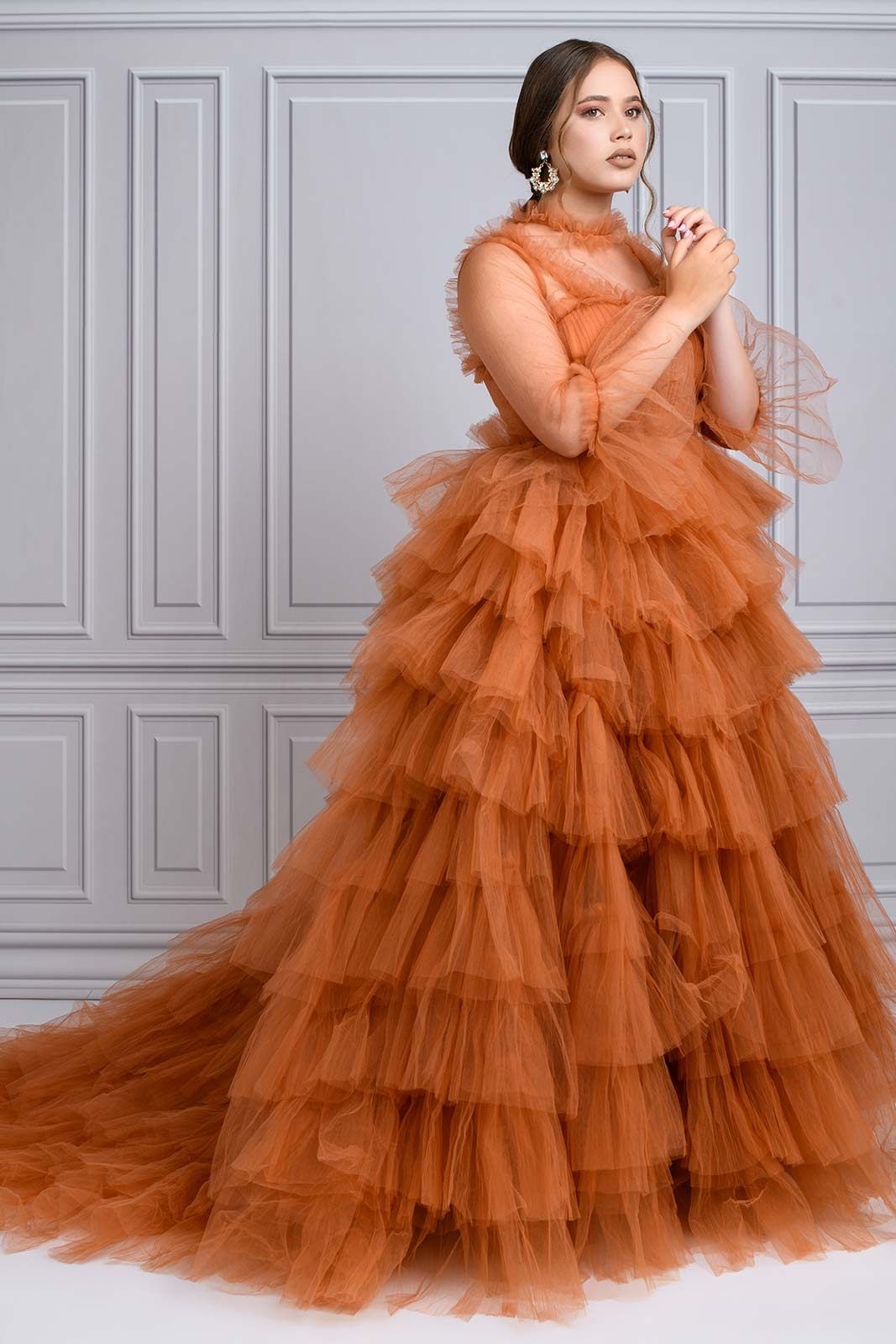 A stunning burnt orange tulle ball gown with tiered ruffles and sheer long sleeves against a gray paneled wall.