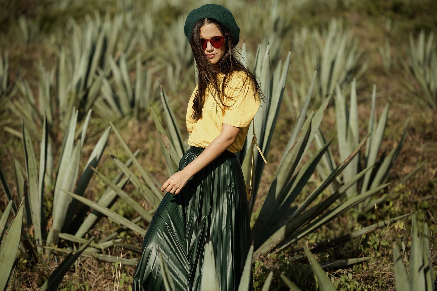 Person in yellow top and sunglasses poses among agave plants in desert landscape.