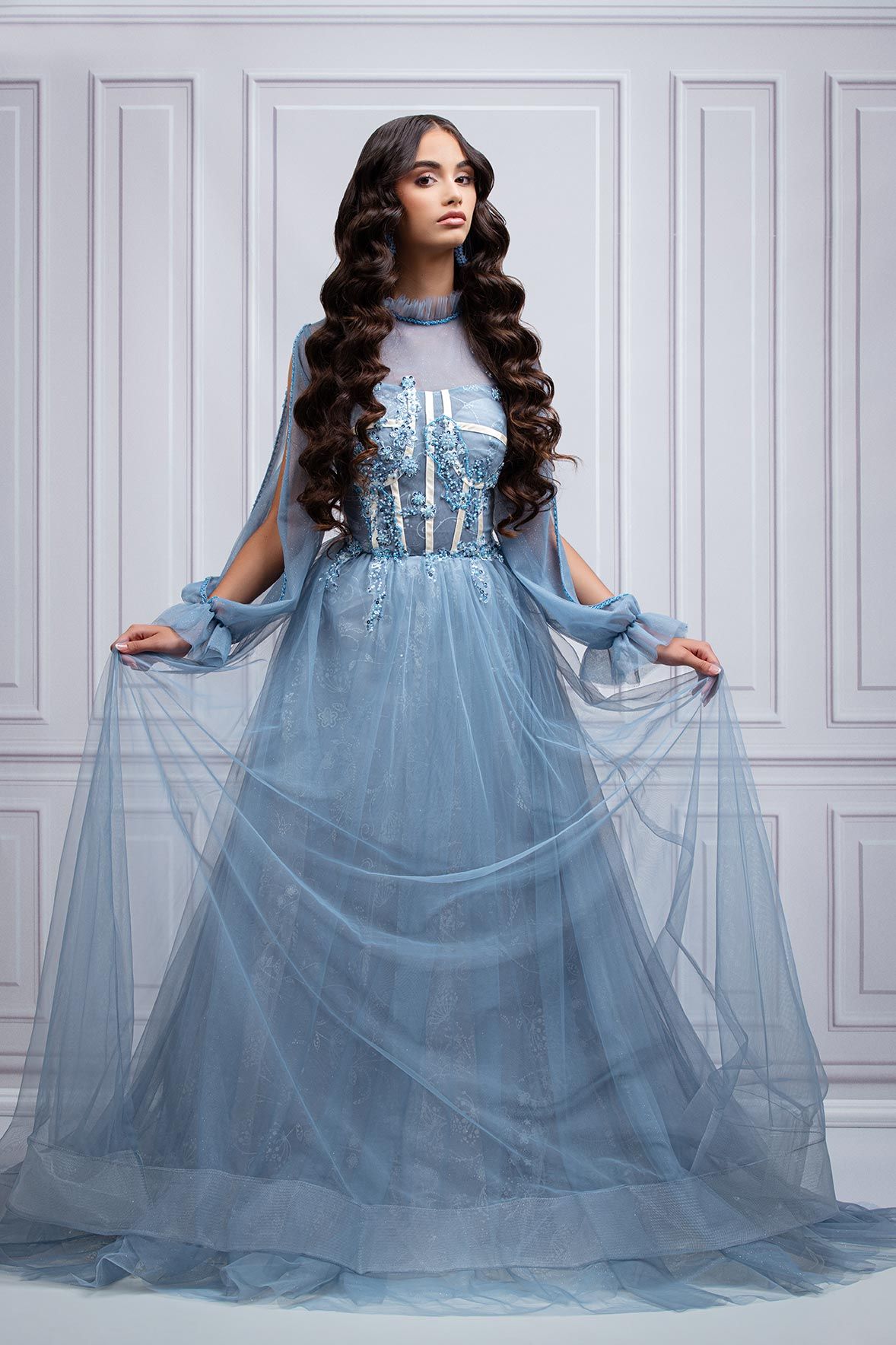 Elegant powder blue tulle gown with long flowing sheer sleeves and embellished bodice against classic white paneled wall.