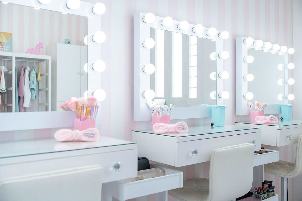 Professional makeup vanity station with illuminated mirrors, white furniture, and pink decorative accents in a bright salon setting.