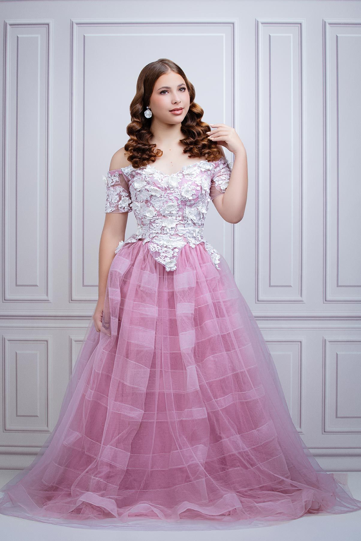 Romantic pink tulle ball gown with lace bodice and off-shoulder sleeves against classic white wall backdrop.