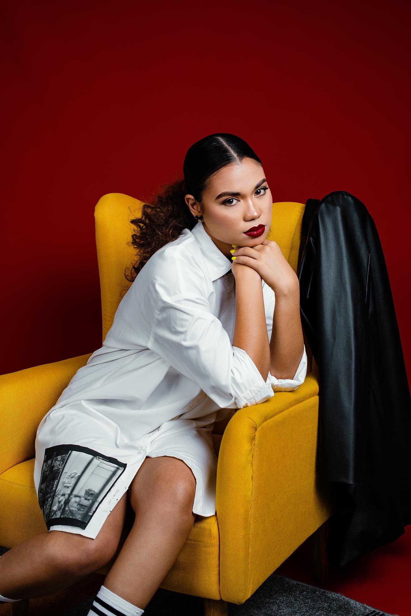 Fashion model in white shirt dress poses on yellow armchair against red background with dramatic styling.