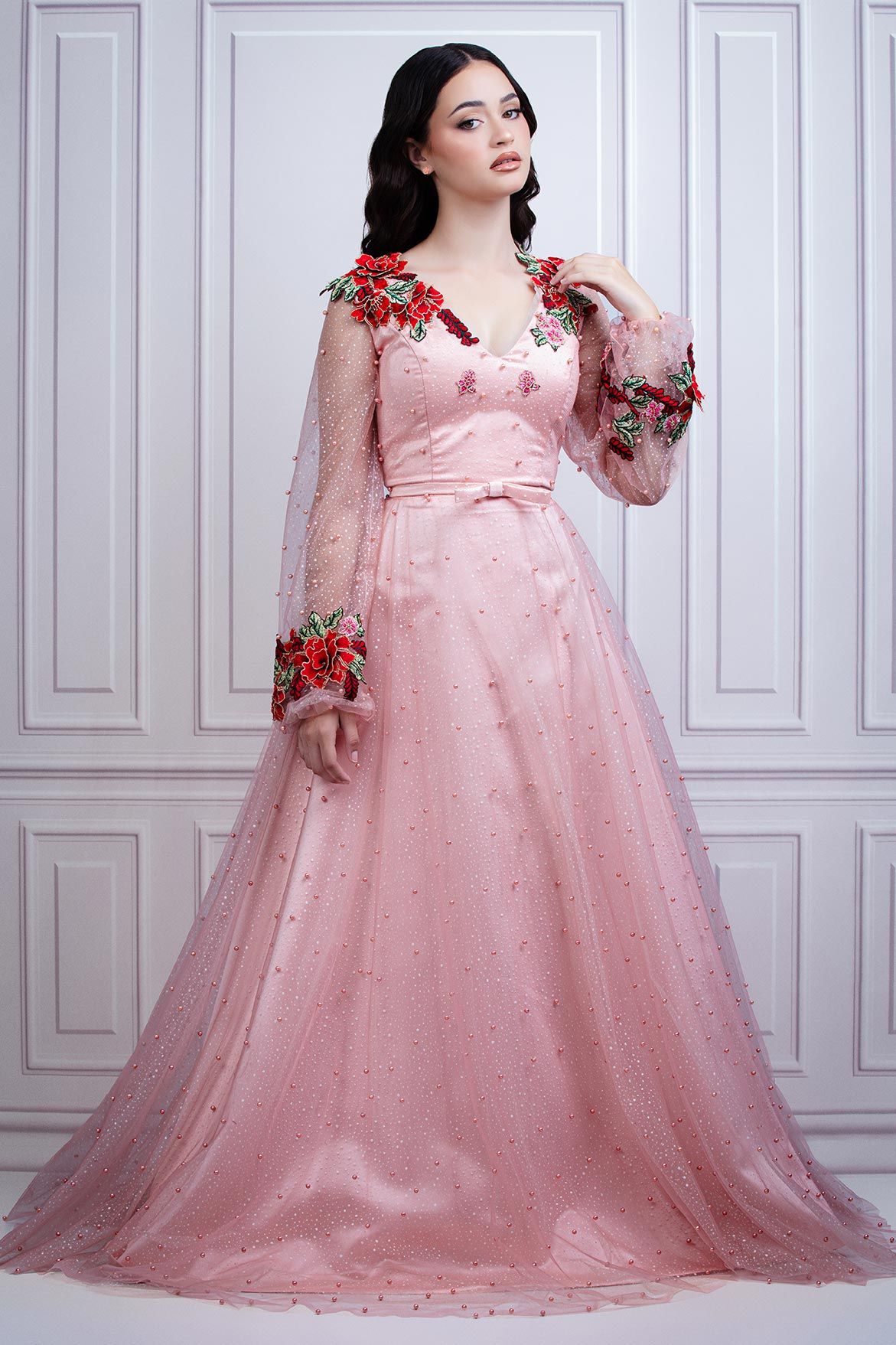 Pink tulle gown with red floral appliques and sheer long sleeves photographed against white wall.