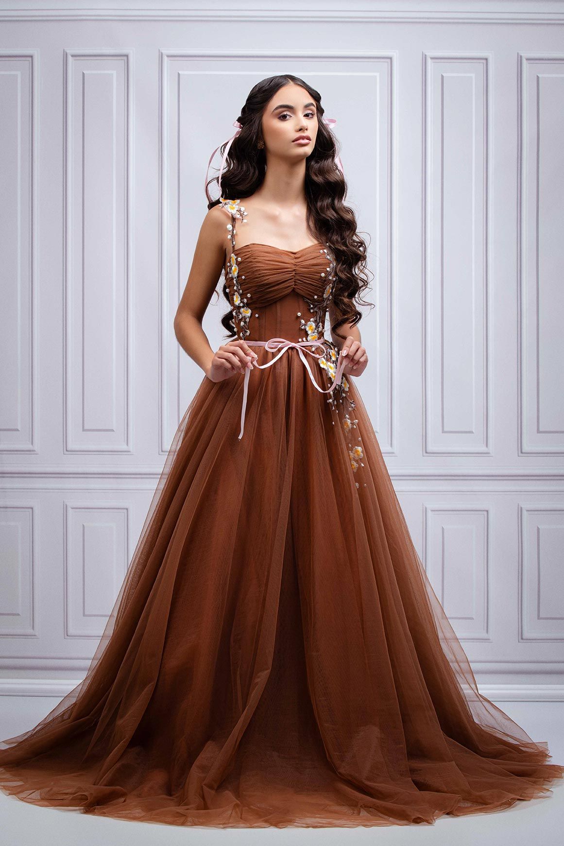 Elegant brown tulle ball gown with corset bodice and floral appliques stands against a classic white paneled wall.