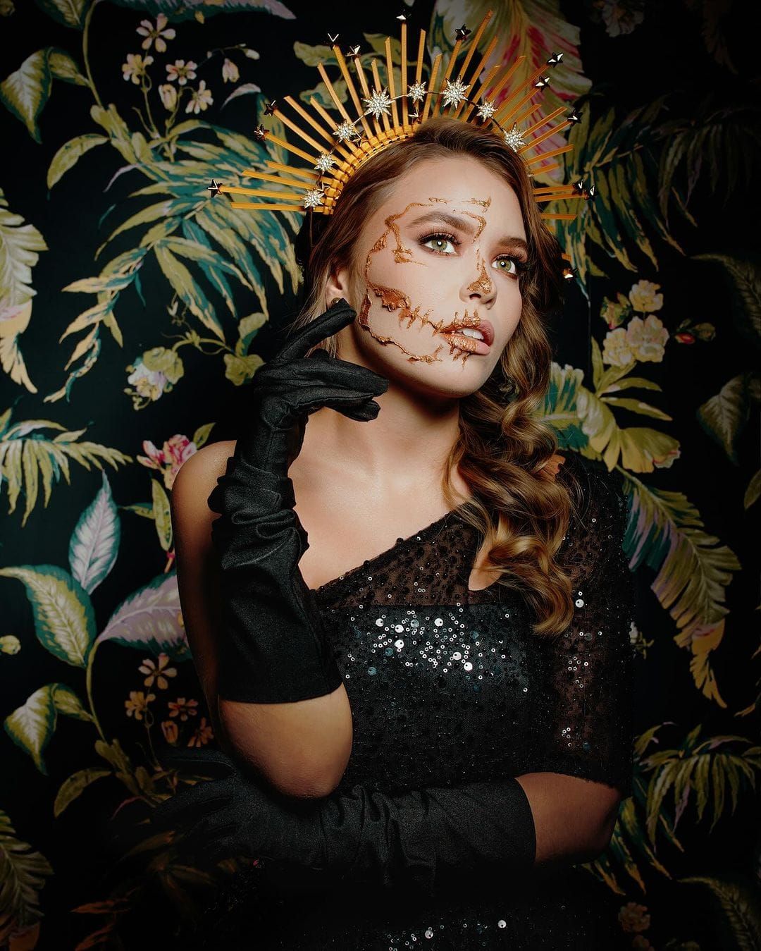 Artistic Halloween portrait against floral wallpaper showing golden crown, dark gloves, and intricate face paint design.