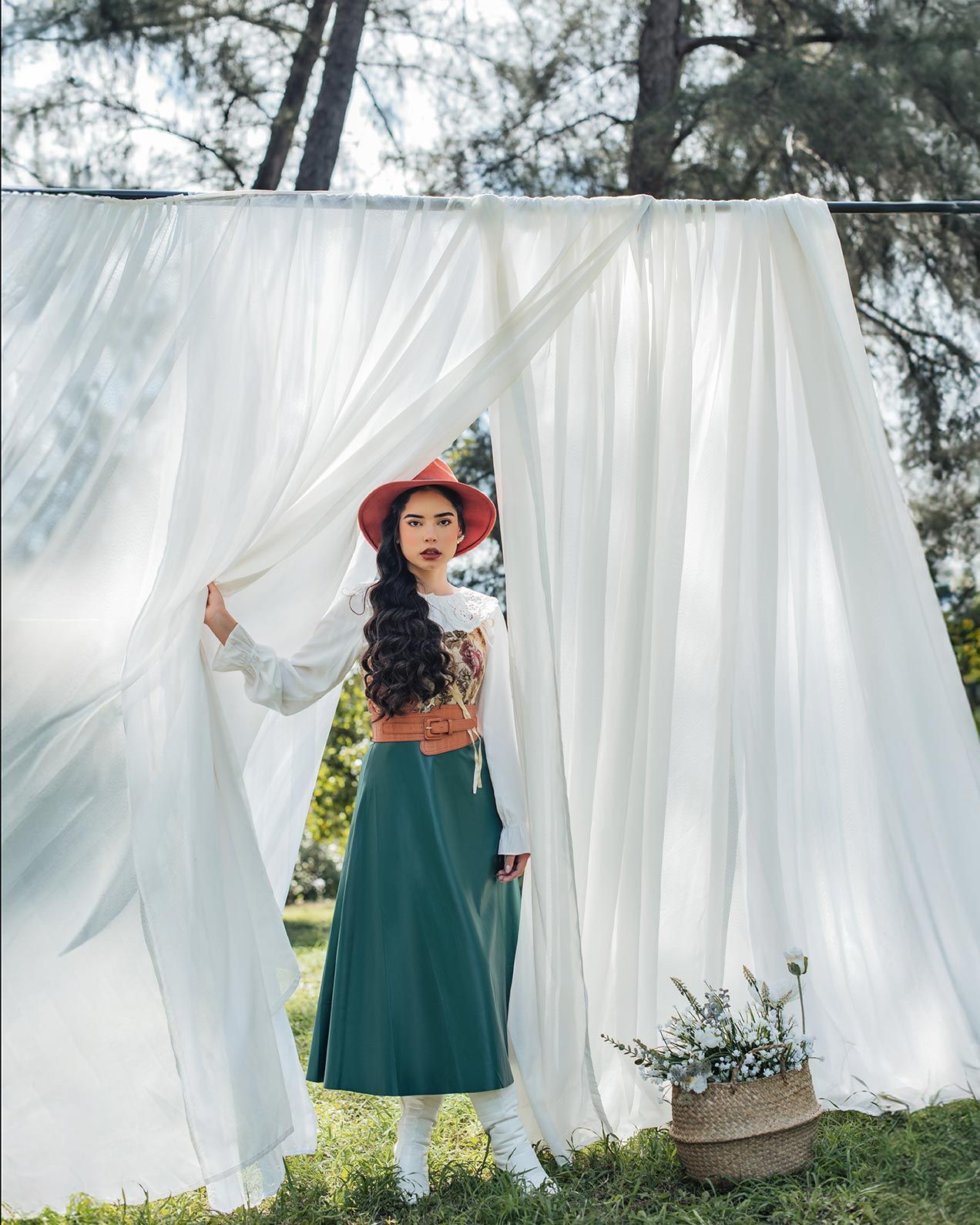 A figure stands against white flowing curtains outdoors wearing a long green skirt, white blouse and red hat in bohemian style.