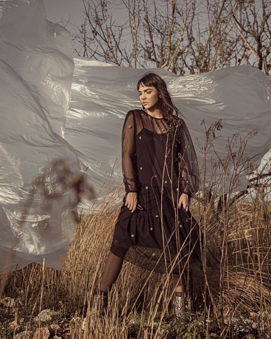 Fashion portrait of person in sheer black dress standing in tall autumn grass against blurred white backdrop.