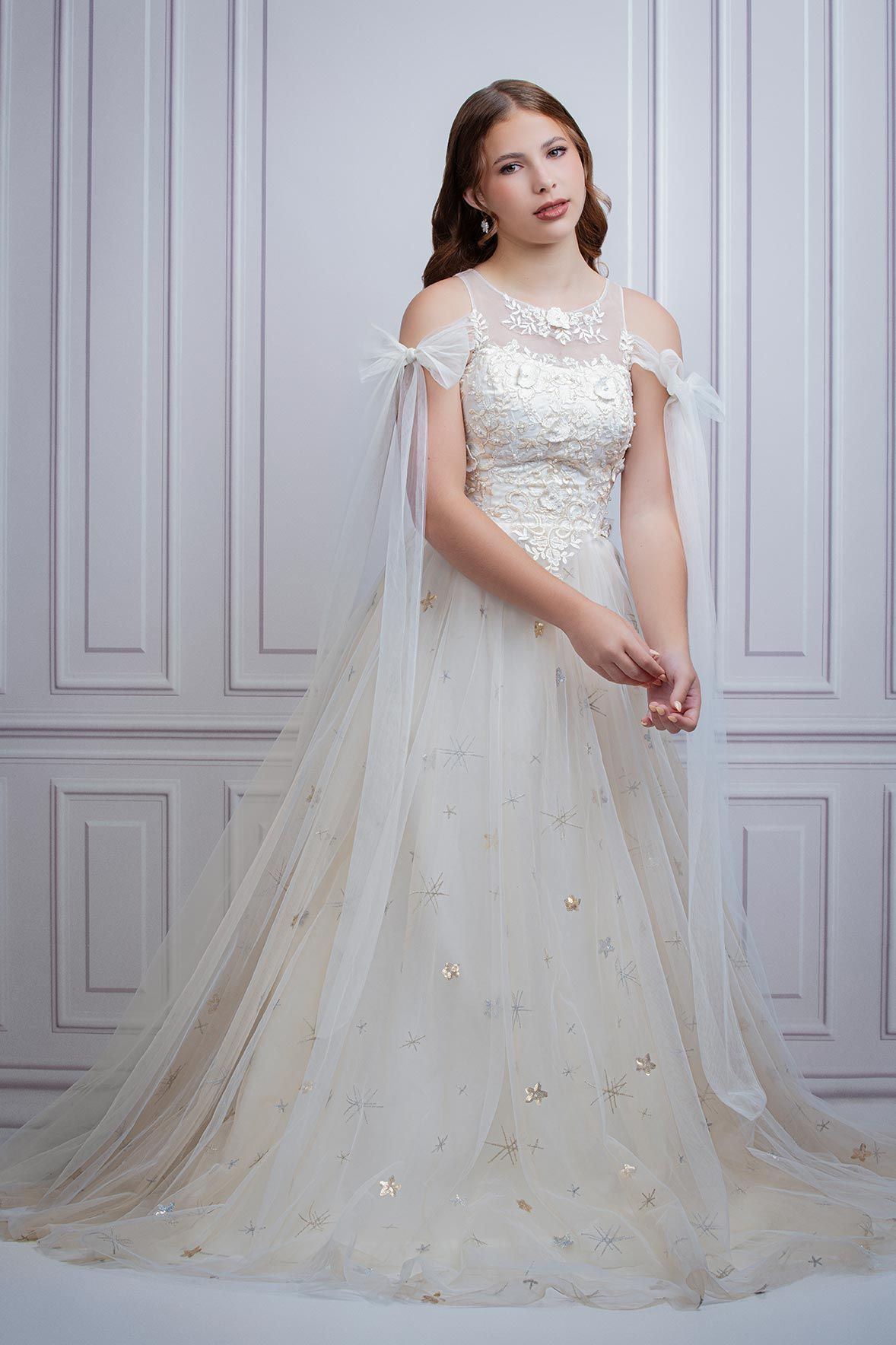 A romantic off-shoulder tulle wedding gown with delicate floral details and flowing cape stands elegantly against white walls.
