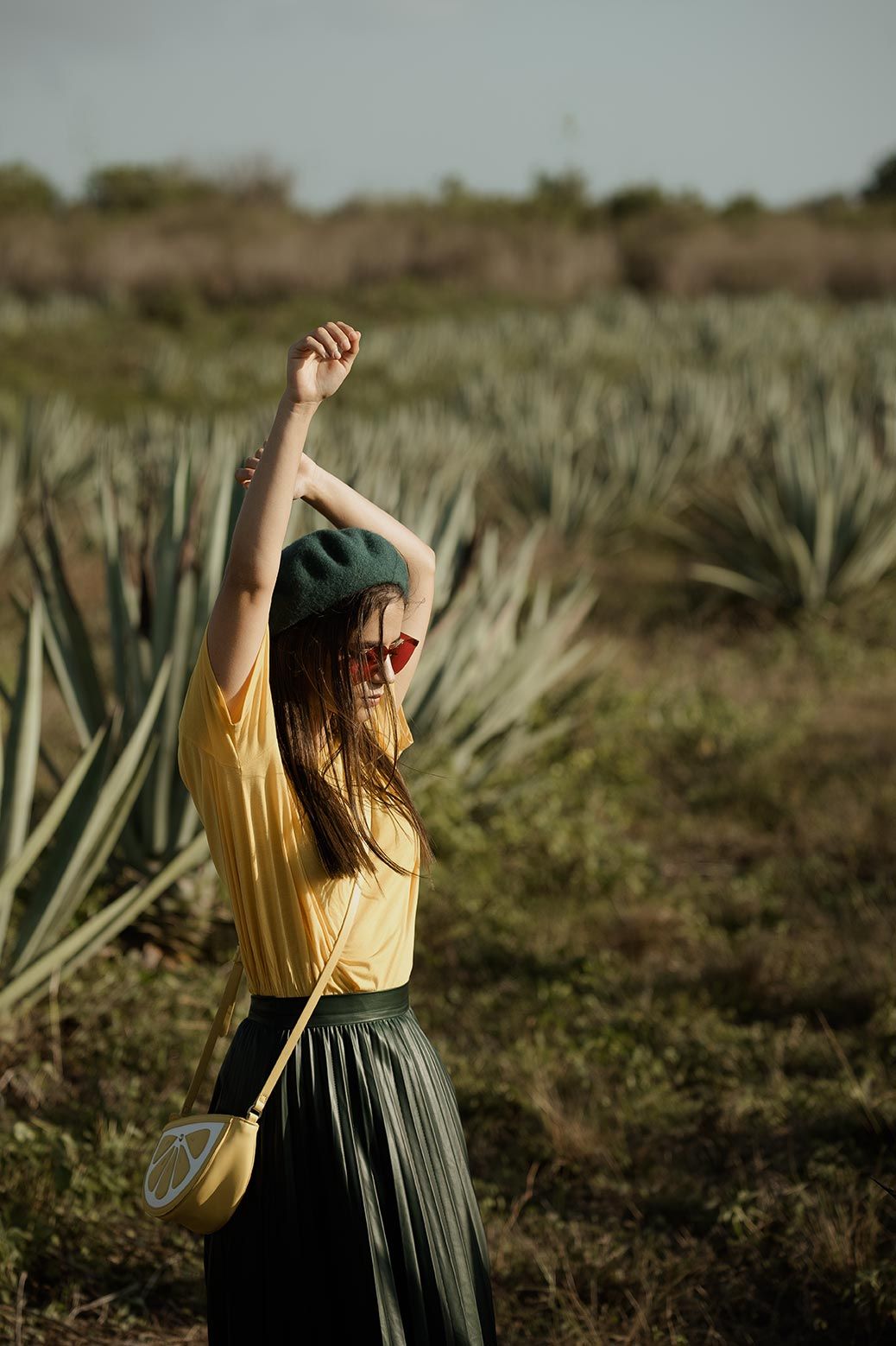 Person in a yellow shirt and dark skirt dancing joyfully in a field of agave plants at sunset.
