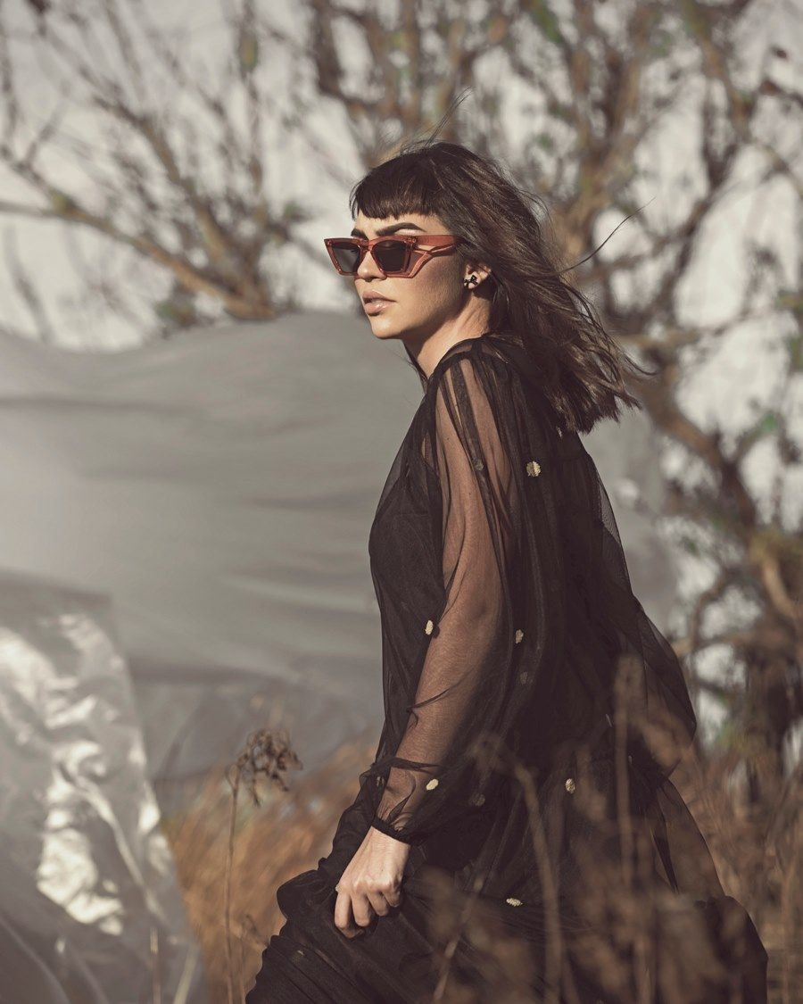 Person in dark sheer coat and sunglasses poses dramatically against bare winter trees in moody outdoor fashion photo.