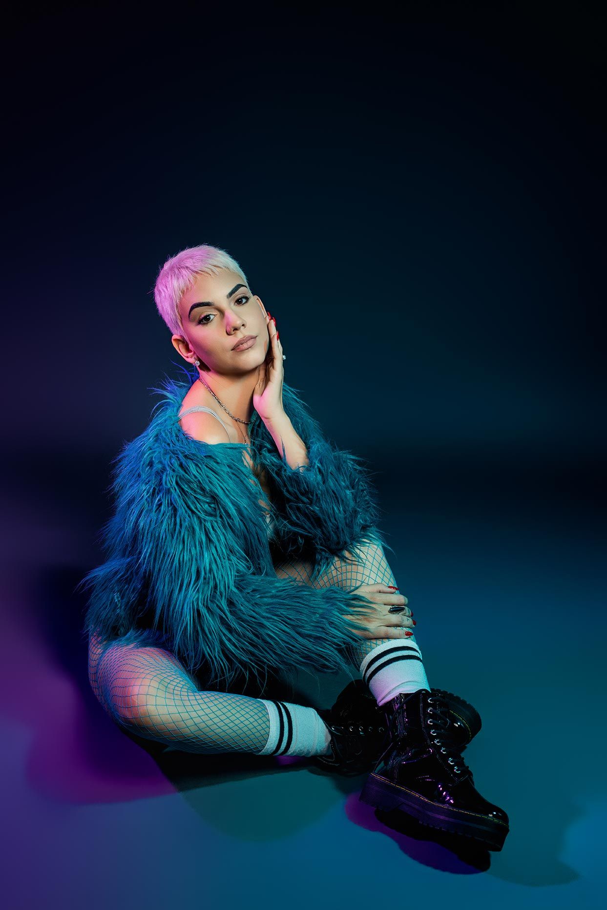 Person in teal faux fur coat and platform boots poses stylishly against dark background with purple and blue lighting.