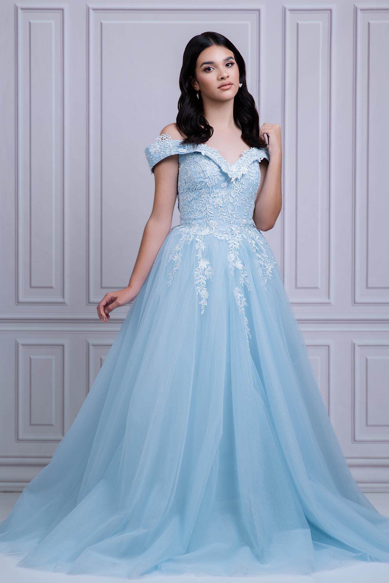 Elegant light blue tulle evening gown with off-shoulder design and delicate lace appliques against white paneled wall.