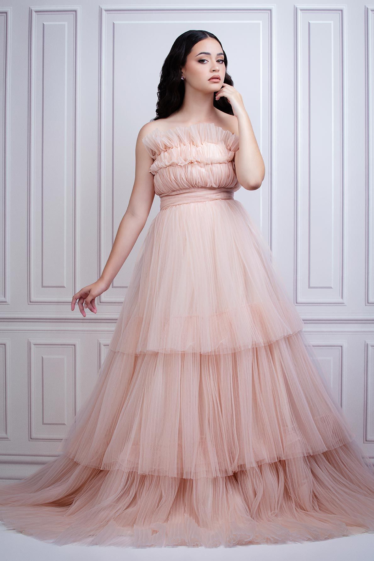Elegant blush pink layered tulle gown with strapless ruched bodice and flowing tiered skirt photographed against white paneling.