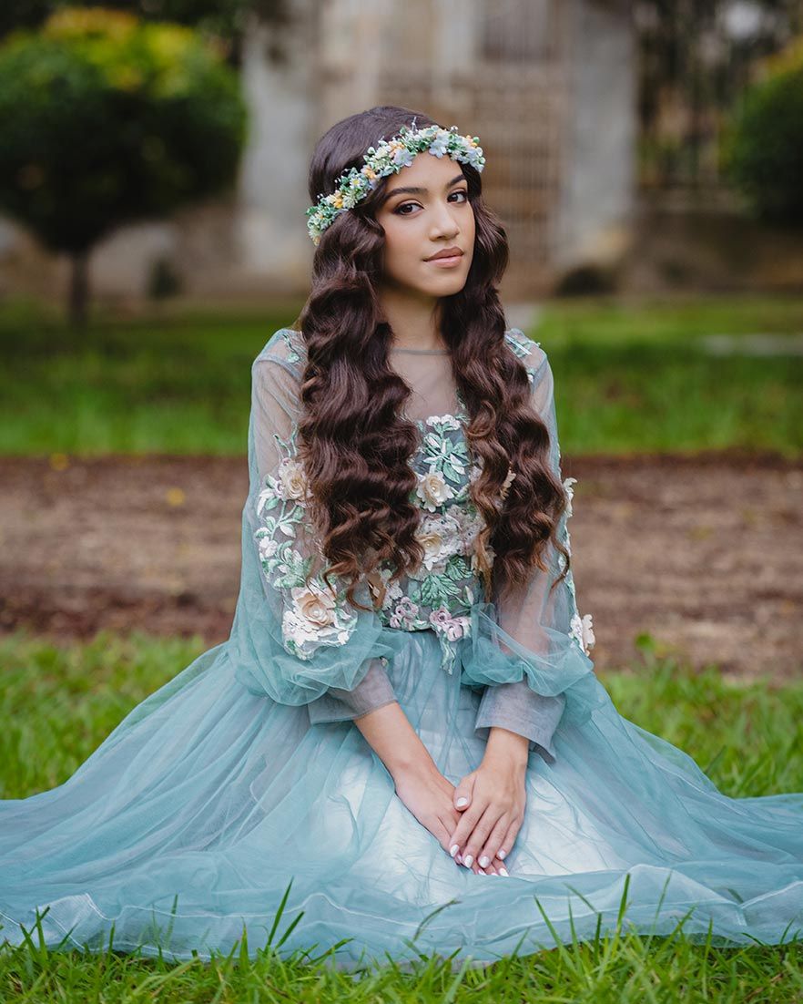 A flowy mint blue dress with delicate lace details and flower crown adorns someone sitting in a dreamy outdoor garden setting.
