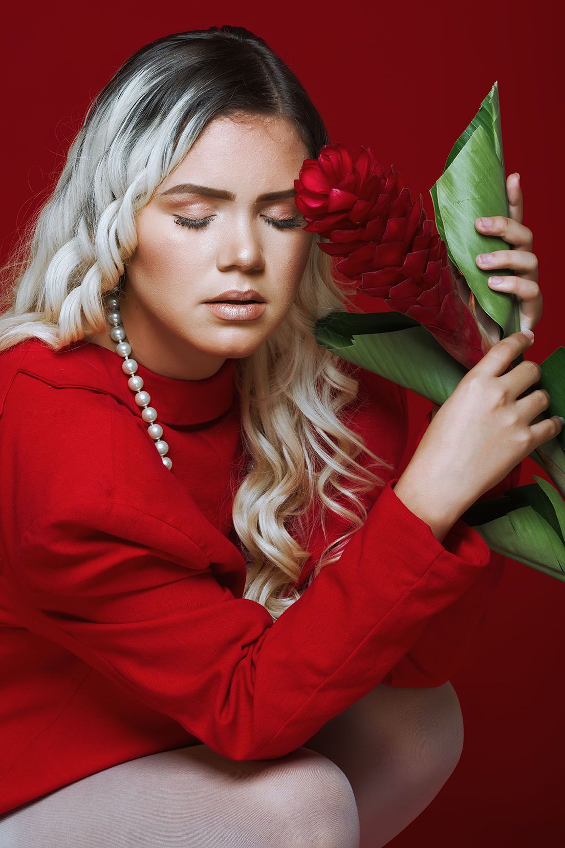 Fashion editorial portrait with red rose, featuring platinum blonde hair and red sweater against burgundy background.