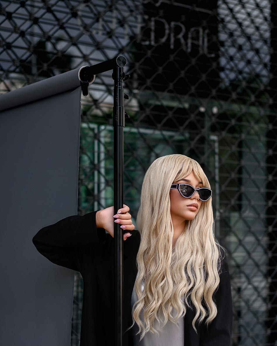 A figure in sunglasses with long wavy platinum blonde hair poses against an urban backdrop with geometric patterns.