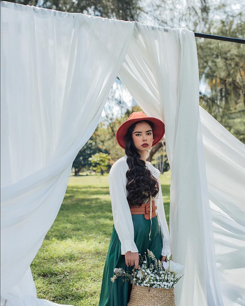 A romantic fashion scene with white curtains, red hat, emerald skirt and delicate wildflower bouquet in a garden setting.