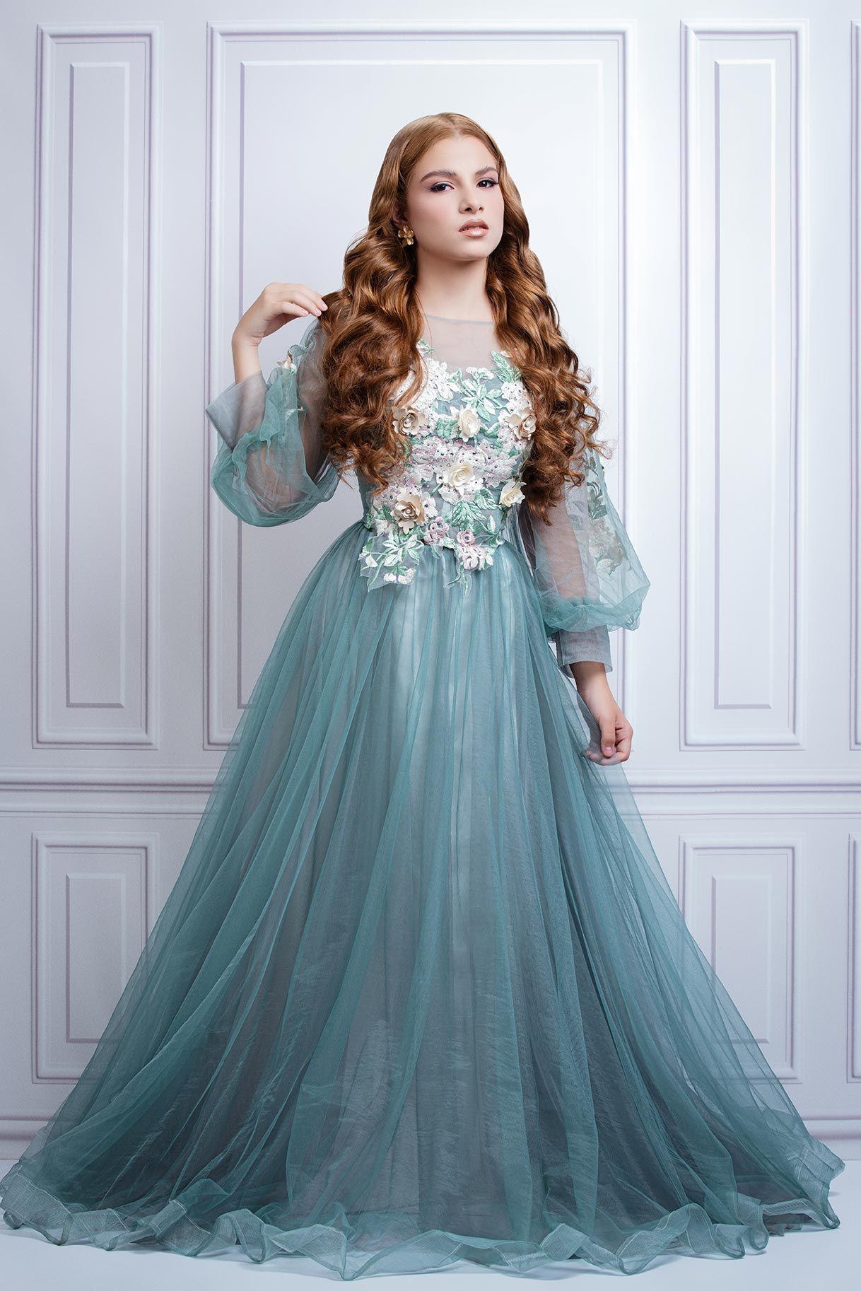 Ethereal aqua blue tulle gown with floral lace appliques and billowing sheer sleeves against white paneled walls.