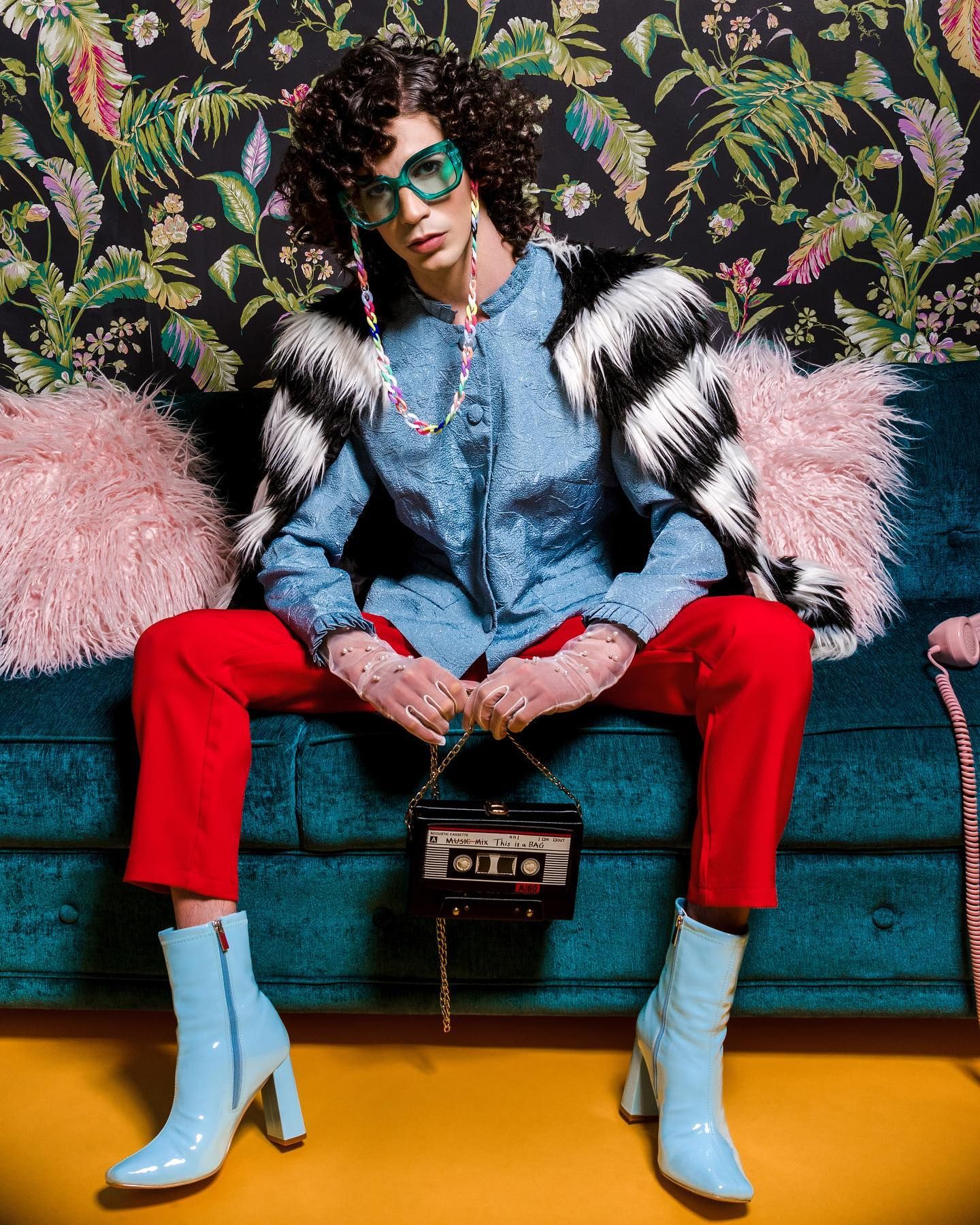 Fashion portrait with denim jacket, red pants, light blue boots and floral wallpaper against teal velvet couch.