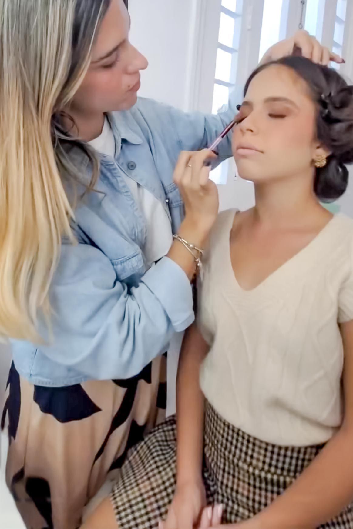 A makeup artist in a denim shirt applies cosmetics to a client wearing a beige sweater and plaid skirt.