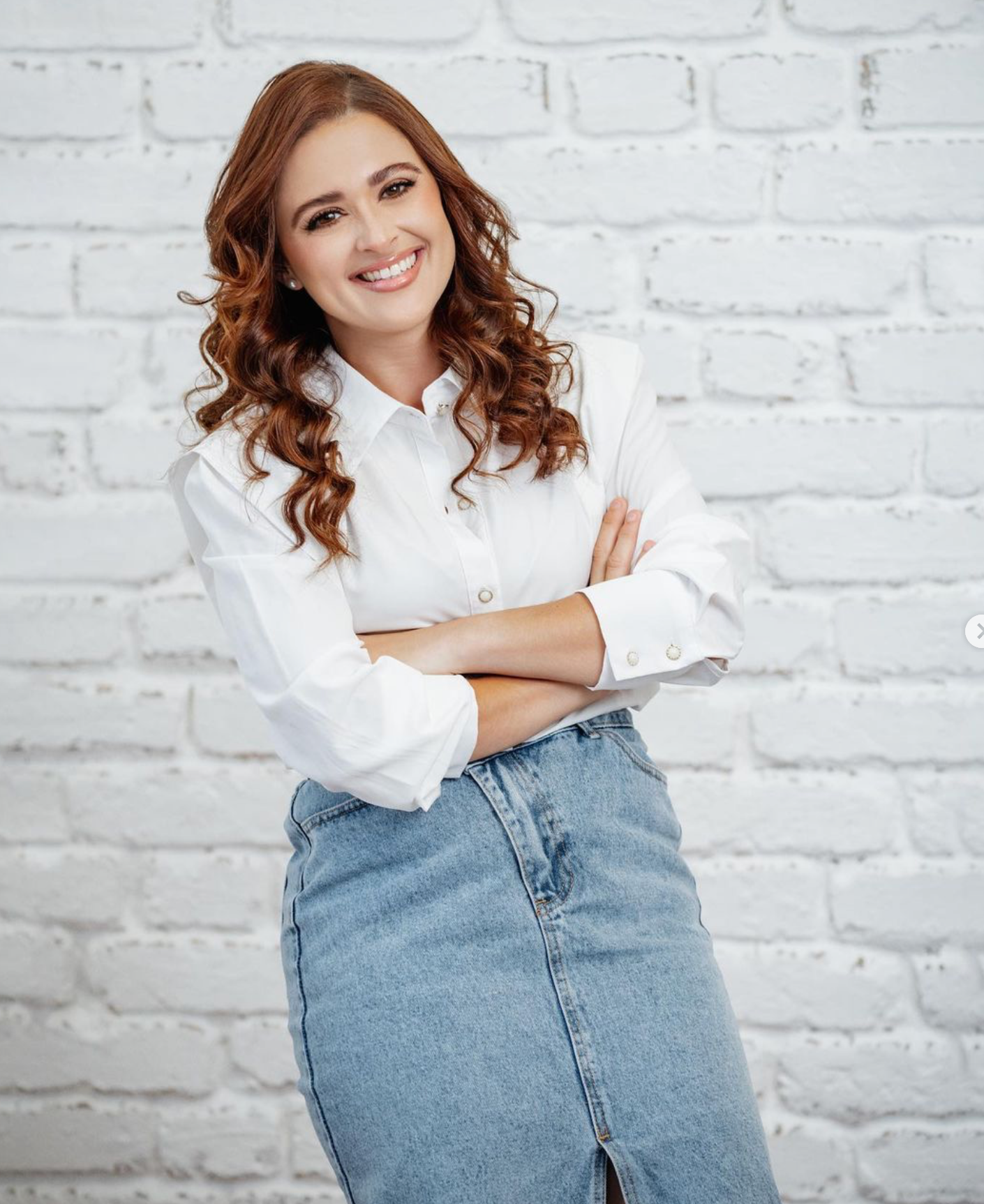 Woman in white button-up shirt and denim skirt smiling while standing against white brick wall.