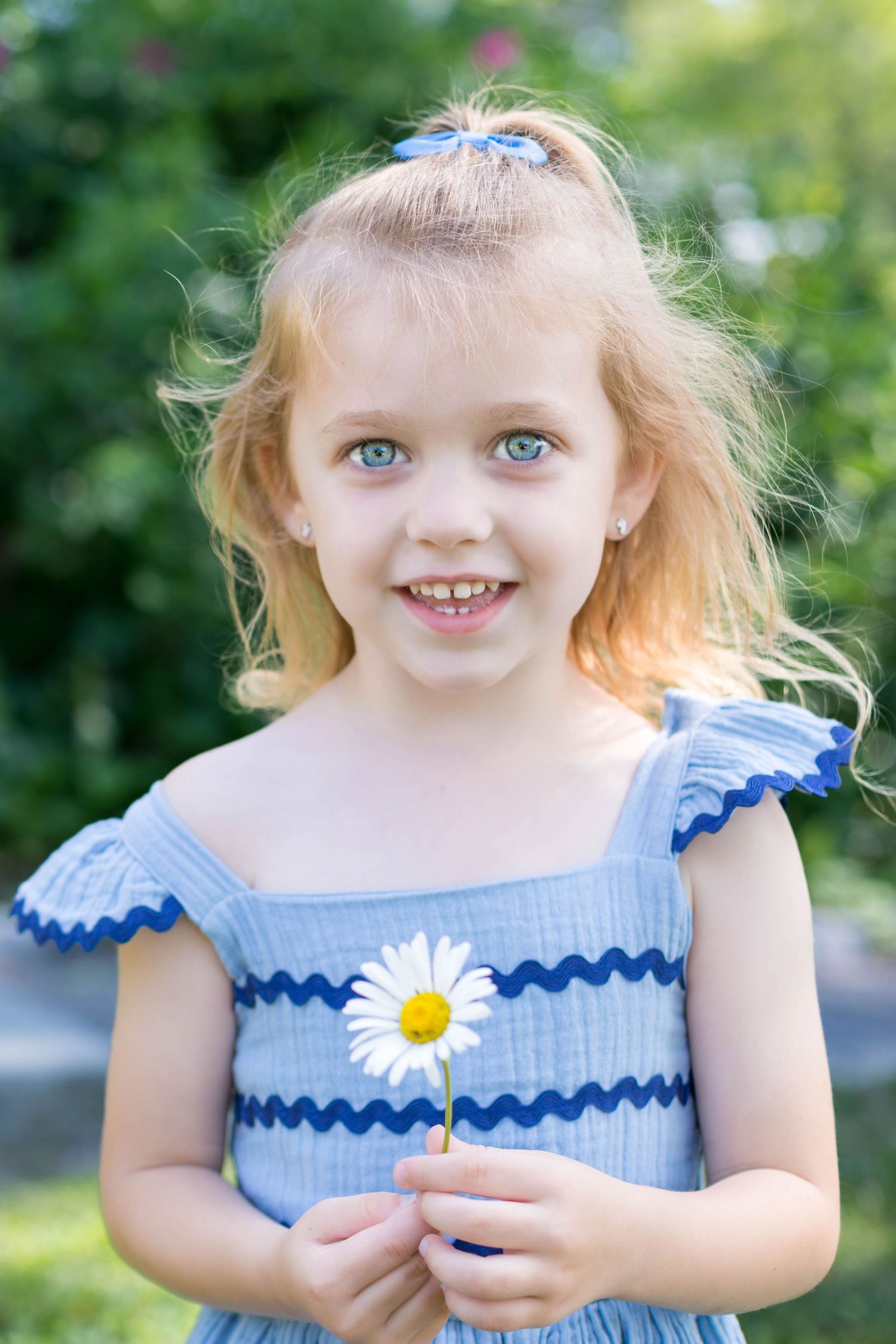 Young girl in a blue dress smiles while holding a daisy.