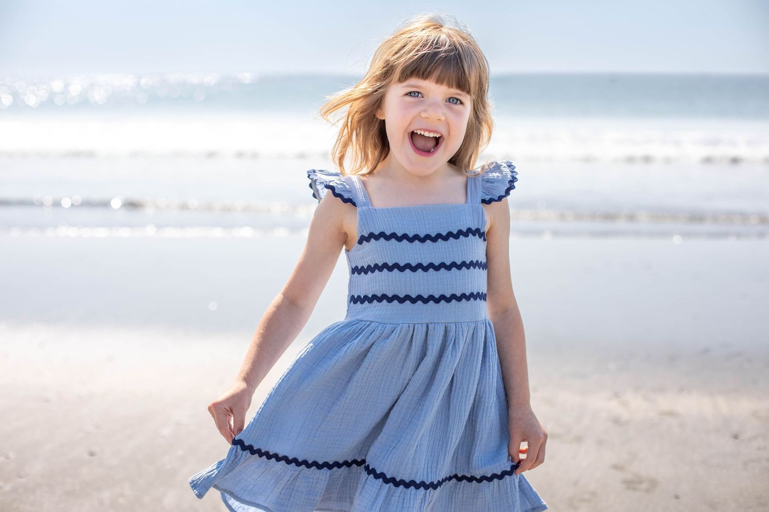 Little girl standing on the beach, holding her blue dress out to the side as her brown hair blows in the wind.