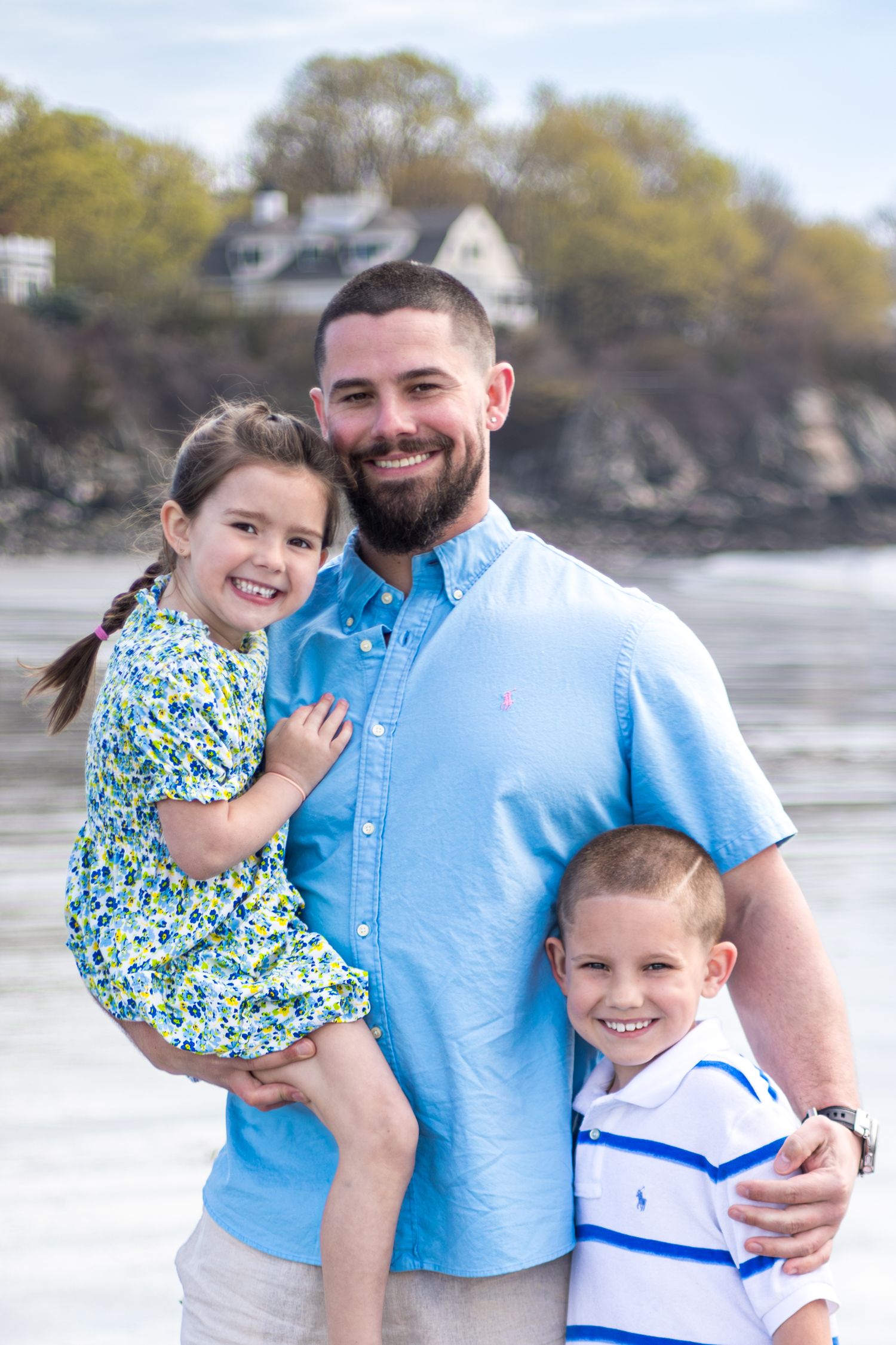 Father holding his daughter and hugging his son at the beach, all smiling at the camera.