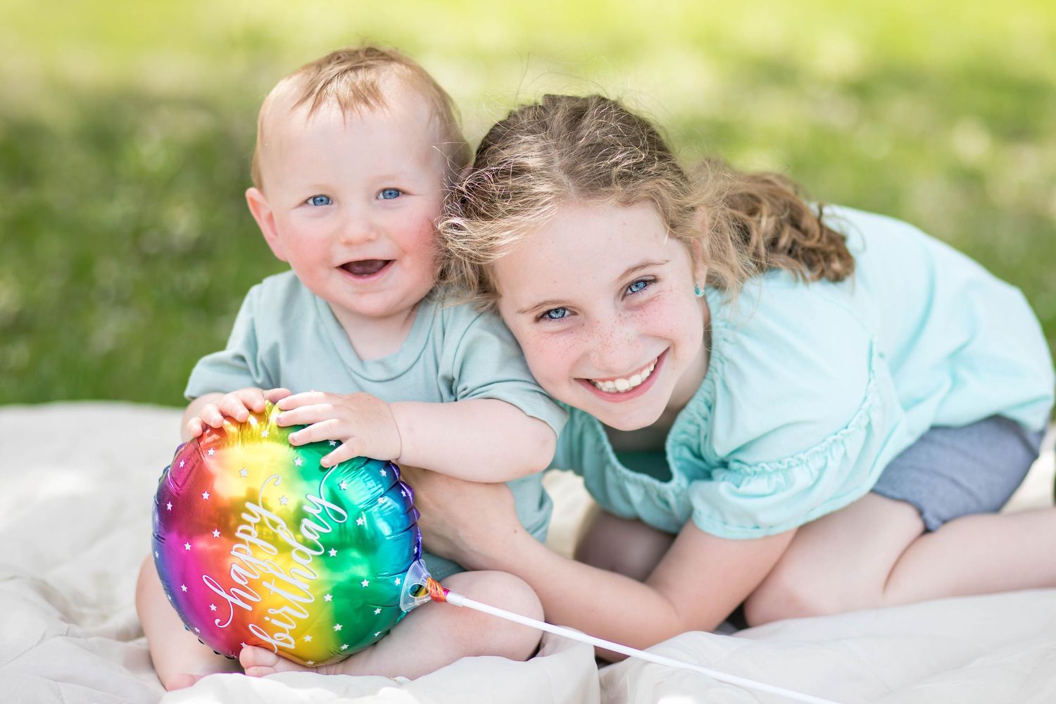 Big sister snuggles her one-year-old brother, who is holding a rainbow birthday balloon outside on a blanket.