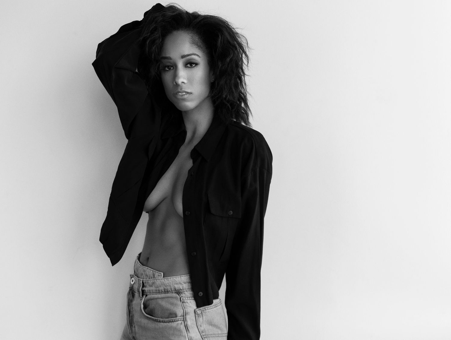 Black and white portrait of a woman in an open black jacket and jeans posing confidently against a white background.