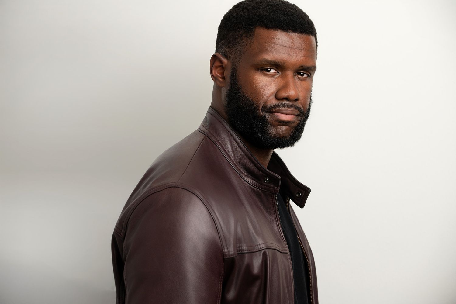 A confident man in a dark brown leather jacket poses against a light gray background with a serious expression.