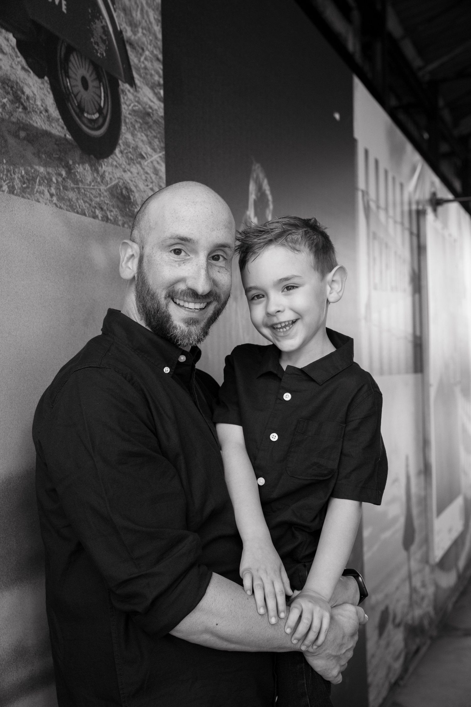 Black and white photo of a bald bearded man holding a smiling young boy, both wearing black shirts.