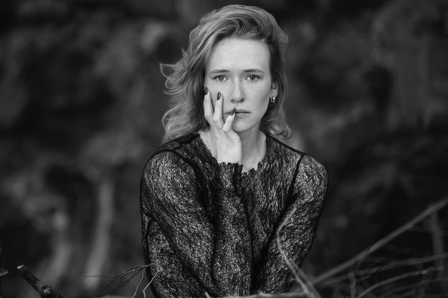 Black and white portrait of a young woman in lace top, hand near face, with blurred nature background.