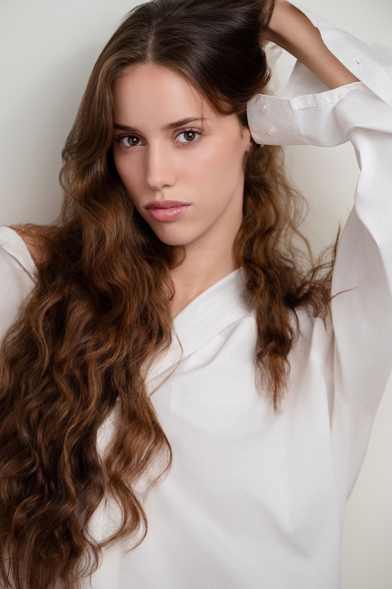 Young woman with long wavy brown hair wearing a white oversized shirt, posing against a light background.
