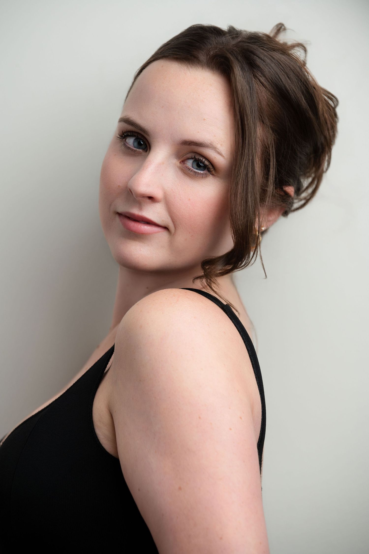 Young woman with updo hairstyle wearing black tank top, smiling over shoulder against light gray background.